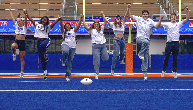 Seven Boise State fans wearing white apparel and jumping on the Blue