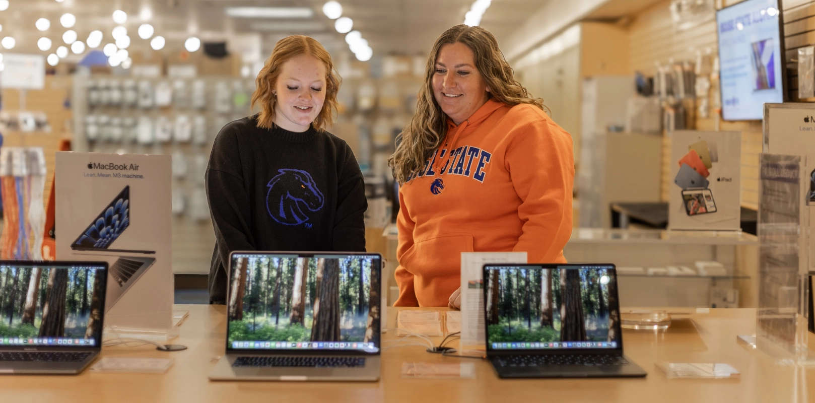 Two women in Bronco Gear sitting on the grass, smiling. Welcome to the Boise State University Bronco Shop!