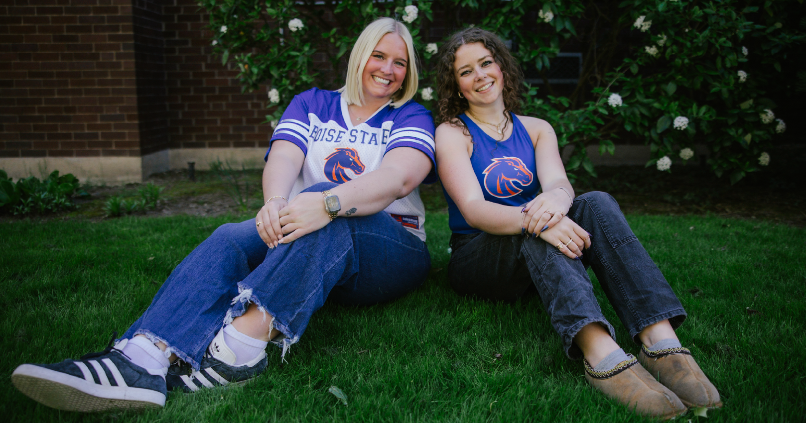 Large image of two women in Bronco Gear sitting on the grass, smiling, Welcome to the Boise State University Bronco Shop!