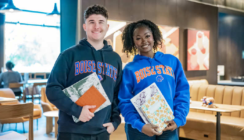 Two Boise State students wearing Broncos hoodies and holding colorful notebooks and folders